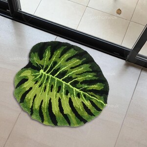 May include: A green and black leaf-shaped rug. The rug features a detailed design mimicking the veins and patterns of a monstera leaf. The rug is placed on a tiled floor near a glass door.