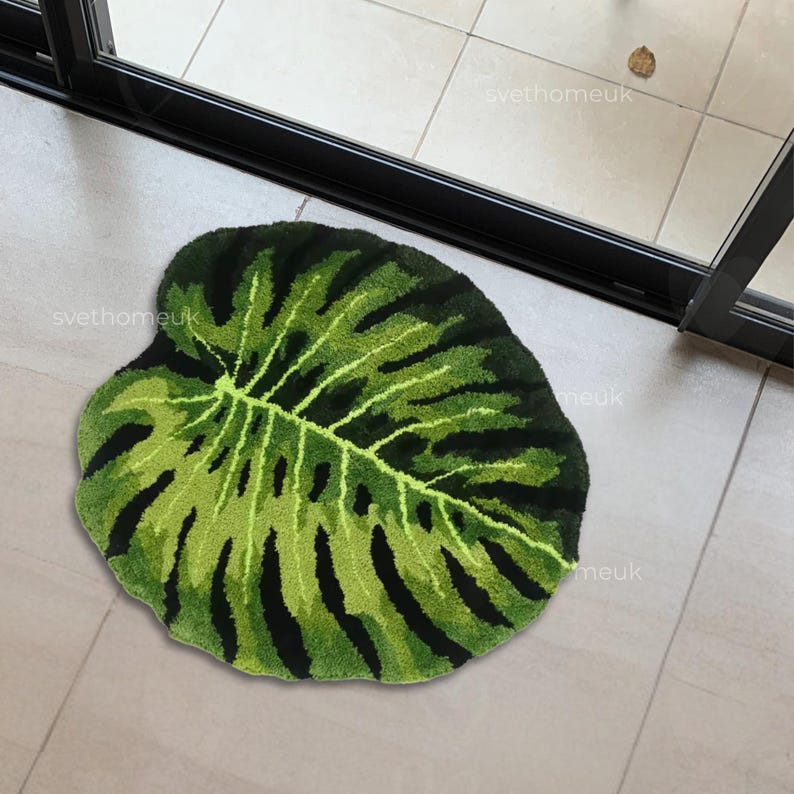 May include: A green and black leaf-shaped rug. The rug features a detailed design mimicking the veins and patterns of a monstera leaf. The rug is placed on a tiled floor near a glass door.
