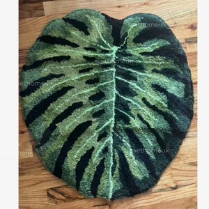 May include: A green and black leaf-shaped rug. The rug is designed to resemble a Monstera plant leaf, with a detailed pattern of veins and dark green and black sections. The rug is placed on a wooden floor.