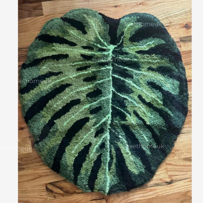 May include: A green and black leaf-shaped rug. The rug is designed to resemble a Monstera plant leaf, with a detailed pattern of veins and dark green and black sections. The rug is placed on a wooden floor.
