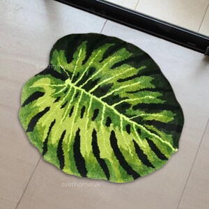 May include: A green and black leaf-shaped rug. The rug is designed to resemble a Monstera plant leaf, with a detailed pattern of veins and cutouts. The rug is made of a soft, plush material and is suitable for indoor use.