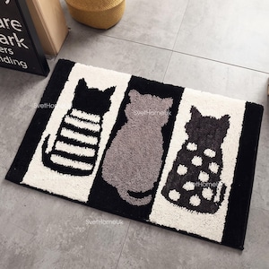 May include: A rectangular bath mat featuring three cat silhouettes in a row. The left cat is black with white horizontal stripes, the middle cat is solid gray, and the right cat is black with white polka dots, all set against a white and black background.