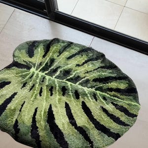 May include: A leaf-shaped rug with a green and black pattern. The rug has a textured surface and a light green center with dark green and black stripes radiating outwards. The rug is on a light-colored tile floor.