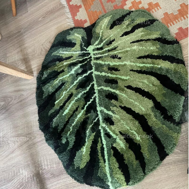 May include: A leaf-shaped rug in shades of green and black, resembling a Monstera plant leaf. The rug features a textured, tufted design with a central vein and dark stripes, creating a naturalistic look. The rug is placed on a wooden floor.