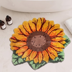 Könnte beinhalten: Ein getufteter Teppich in Sonnenblumenform mit einer dunkelbraunen Mitte, leuchtend gelben und orangen Blütenblättern sowie grünen Blättern an der Basis. Der Teppich hat eine strukturierte, flauschige Oberfläche und liegt auf einem hellen Boden.
