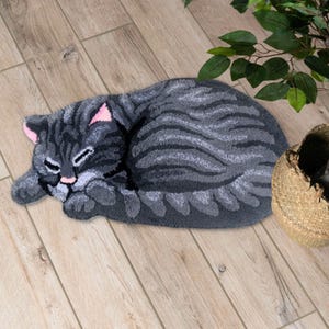 May include: A grey cat-shaped bath mat with a fluffy texture. The cat is curled up in a sleeping position with its eyes closed.