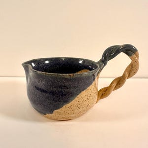 May include: Handmade ceramic pitcher with a twisted handle. The pitcher has a two-tone design, with a dark blue upper section and a speckled tan lower section. The handle is a twisted rope design, transitioning from dark blue to tan.