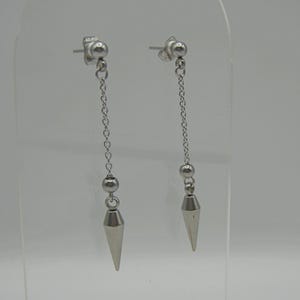 May include: A pair of silver-colored drop earrings. Each earring features a small ball stud, a chain, a small ball, and a cone-shaped pendant. The earrings are displayed against a light gray background.