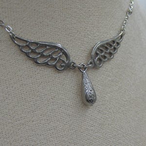 May include: Silver necklace with a teardrop pendant and two angel wings. The wings are open and the pendant hangs from the center.