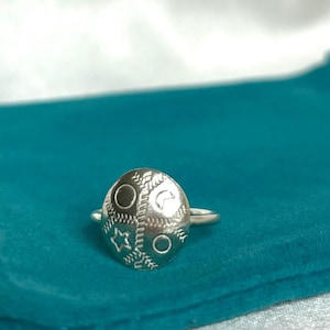 May include: A silver ring with a round, textured top featuring a geometric design. The ring is on a teal velvet pouch.