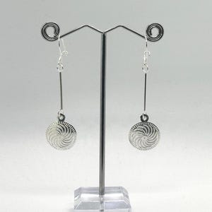May include: A pair of silver-toned dangle earrings. Each earring features a long, thin bar suspended from a hook, with a circular pendant at the bottom. The pendant has a spiral design.