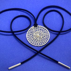 Puede incluir: Un collar con un colgante circular de plata, con un diseño intrincado, que cuelga de un cordón negro con puntas plateadas. El colgante está hecho de alambre delicado e entrelazado, creando un efecto de encaje.