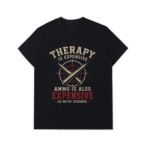 May include: A black short-sleeved t-shirt featuring a graphic with two crossed bullets over a red and white target reticle. The text reads: THERAPY IS EXPENSIVE AMMO IS ALSO EXPENSIVE SO WE'RE SCREWED. The design uses white, red, and beige colours.