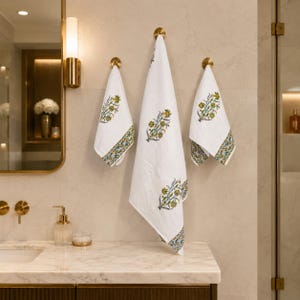 May include: Three white cotton towels hanging on brass hooks. Each towel features a repeating floral block print pattern in shades of green and yellow, with a decorative border along the bottom edge. The fabric has a visible textured weave.