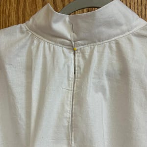 May include: Close-up of a white garment with a high collar, hanging on a gray hanger. The collar is a lighter shade of white than the rest of the garment. A yellow pin secures the collar's closure. The fabric appears to be linen or a similar lightweight material.