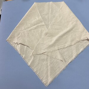 May include: A triangular scarf made of light beige, textured fabric with a visible weave. The edges of the scarf are unfinished and feature a frayed, raw fringe detail along the entire perimeter, creating a simple and unadorned appearance.