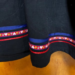 May include: A black skirt featuring horizontal decorative trim near the hem. The trim consists of a solid navy blue ribbon, a red ribbon with a repeating pattern of white hearts and small yellow and blue flowers, and a thin red ribbon below.