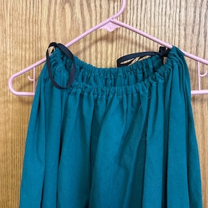 May include: A solid teal-colored garment featuring a gathered neckline with a drawstring closure. The neckline is secured with thin black ribbon ties that are knotted at the top. The fabric appears opaque and drapes downward in vertical folds.