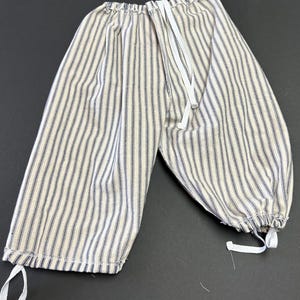 May include: A pair of loose-fitting trousers featuring a vertical pinstripe pattern in dark blue and off-white. The garment includes an elasticized waistband and adjustable white fabric ties at the bottom of each leg opening for a gathered fit.