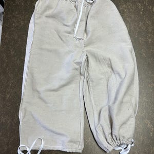 May include: Off-white, textured trousers featuring an elasticized waistband with a central white drawstring tie. The pants have a loose, wide-leg silhouette with gathered, adjustable cuffs at the bottom of each leg, secured by white drawstring ties.