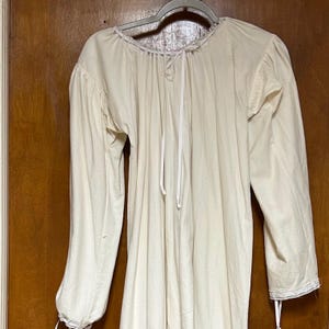 May include: An off-white long-sleeved shirt featuring a gathered neckline with a thin drawstring tie. The garment has full sleeves that taper to cuffs with delicate white lace trim and additional thin ties at the wrists for a secure fit.