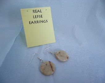May include: A pair of earrings made from real lefse, a traditional Norwegian flatbread. The earrings are oval shaped and have a light brown color with brown flecks. The earrings are hanging from silver hooks.