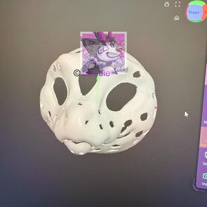 May include: A 3D-modeled white mask with large eye and mouth openings. A small, square image of a cartoon character with pink and purple accents is superimposed on the mask. The image is on a computer screen.