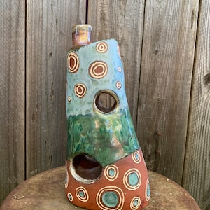 May include: A ceramic vase with an abstract design. The vase is brown, green, and blue with white circles and rings. The vase has a unique shape with cutouts.