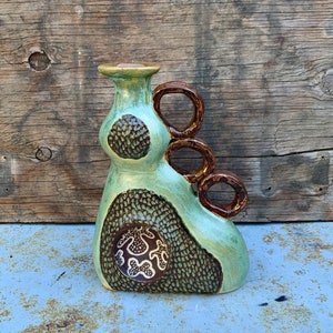 May include: A green and brown ceramic vase with a unique, abstract design. The vase has three loops on the side and a textured surface.