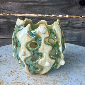 May include: A ceramic vessel with a glossy finish in shades of green and cream, featuring an irregular, organic shape with multiple circular cutouts and small, raised white spherical accents distributed across the textured surface.