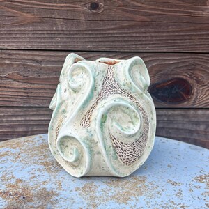 May include: A ceramic vase featuring a light green and cream mottled glaze with raised, swirling sculptural elements. The surface displays a textured, dark brown crackle pattern within the recessed areas of the organic, flowing design.