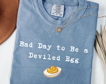 May include: A blue jean Comfort Colors t-shirt with the text "Bad Day to Be a Deviled Egg" and an illustration of a deviled egg. A gold necklace with a pendant is draped across the shirt.