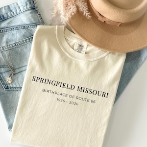 Puede incluir: Camiseta color marfil con el texto negro "SPRINGFIELD MISSOURI BIRTHPLACE OF ROUTE 66 1926-2026". La camiseta se muestra con vaqueros azules, un sombrero beige y un collar.