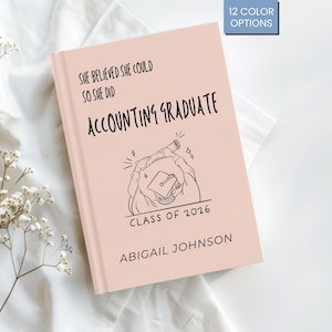Könnte beinhalten: Ein rosa Notizbuch mit dem Text "SHE BELIEVED SHE COULD SO SHE DID ACCOUNTING GRADUATE CLASS OF 2026 ABIGAIL JOHNSON". Das Notizbuch hat eine Illustration zum Thema Abschluss und eine Box mit der Aufschrift "12 COLOR OPTIONS".