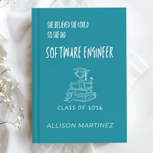 Puede incluir: Un libro de graduación turquesa con texto blanco que dice "She believed she could so she did Software Engineer Class of 2026 Allison Martinez." Una pila de libros con un gorro de graduación y un trofeo está en el centro.