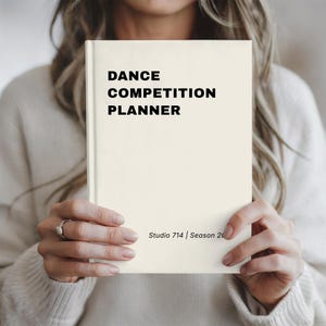 May include: A cream-colored planner with a hard cover featuring the black text DANCE COMPETITION PLANNER at the top and Studio 714 | Season 20 at the bottom. The book is held upright, displaying a clean, minimalist design with bold sans-serif font.