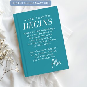 Könnte beinhalten: Ein türkisfarbenes Hardcover-Buch mit weißem Text, der A NEW CHAPTER BEGINS lautet, gefolgt von einer sentimentalen Botschaft über Neuanfänge und Wachstum, die mit dem Namen Alex in einer Schreibschrift endet. Das Buch liegt auf einem weißen Stoff.