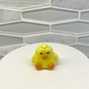 May include: A small yellow chick figurine with orange feet and black eyes. The chick is sitting on a white surface.