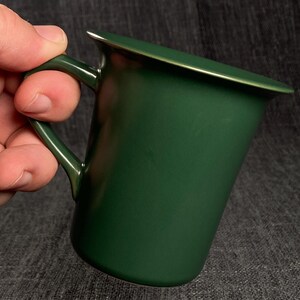 May include: A dark green enamel mug with a curved handle and a silver rim. The mug has a slightly flared top and a smooth, glossy finish. The mug is held by a hand.