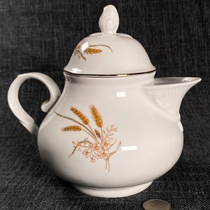 May include: White ceramic creamer with a lid and a handle. The creamer features a wheat and floral design in shades of gold and brown. The rim is trimmed with a gold band. A small coin is next to the creamer.