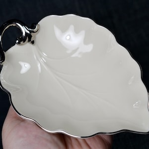 May include: Cream-colored leaf-shaped dish with a silver rim and handle. The dish has a delicate leaf vein pattern and a scalloped edge. The handle is also silver. This decorative dish is likely for serving or display.