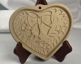 May include: A beige heart-shaped cookie cutter with a detailed embossed design of a bow, oak leaves, and acorns. The cookie cutter is displayed on a dark brown wooden stand against a white background. The design suggests a fall or autumn theme.
