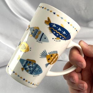 May include: White ceramic mug decorated with stylized fish in shades of blue, yellow, and gold. The fish have geometric patterns and are arranged around the mug. The mug has a white handle and a triangular border.