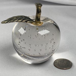 May include: A clear glass apple paperweight with air bubbles. It has a gold-colored stem and a leaf. A quarter is next to the apple for size comparison. The apple is a decorative item.
