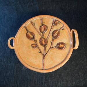 May include: A round, ceramic serving tray with a light orange glaze. The tray features a brown, stylized botanical design in the center and two curved handles. The tray is approximately 10 inches in diameter.