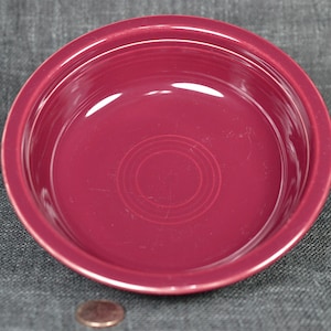 May include: A round, burgundy-colored ceramic bowl with a slightly raised rim. The bowl has concentric circles in the center. The bowl is empty and sits on a gray surface. A penny is in the foreground.