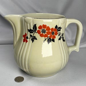 May include: Cream-colored ceramic pitcher with a floral design. The pitcher features a handle and a spout. The floral design includes red flowers and black leaves. The rim of the pitcher has a silver trim.
