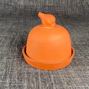 May include: A terracotta butter bell consisting of a dome-shaped lid with a small, pointed handle at the top and a circular base plate. The ceramic material has a matte, earthy orange finish and a smooth, uniform texture throughout the piece.