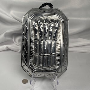 May include: A silver-colored, rectangular mold with a detailed asparagus design. The mold features a bundle of asparagus tied with rope, set within a decorative frame. A black ribbon is attached at the top for hanging.