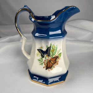 May include: A vintage ceramic pitcher with a blue and white design. The pitcher features a gold-trimmed handle and base, and a butterfly and floral motif. The pitcher is approximately 7 inches tall.